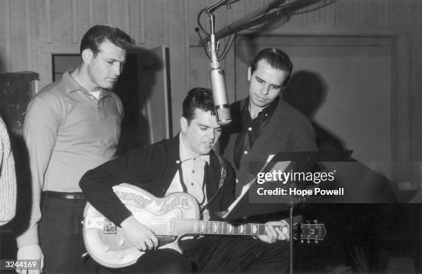 American singer and songwriter Conway Twitty , sits in front of a microphone looking at notes on a music stand while playing a guitar in a recording...