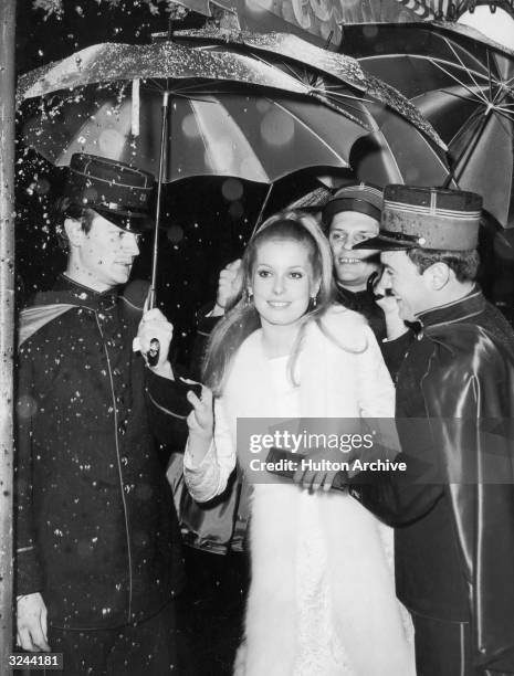 French actor Catherine Deneuve is ushered by gendarmes holding colorful umbrellas to shield her from rain falling from a mechanical device as she...