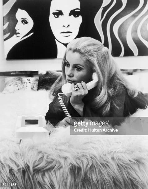 French actor Catherine Deneuve lies on her stomach on a shaggy comforter on top of a bed and holds a telephone receiver up to her ear. There is a pop...