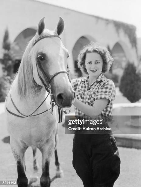 Actor Olivia de Havilland stands next to a horse and holds its reigns in front of cloisters and a fountain. She is wearing jodhpurs and a plaid shirt.