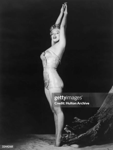 Full-length studio portrait of American actor Marilyn Monroe , wearing a swimsuit, standing on her toes in sand and stretching her arms above her...