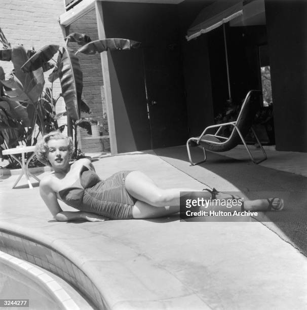 Portrait of American actor Marilyn Monroe lying on a patio next to a swimming pool deck in a strapless bathing suit and high heels.