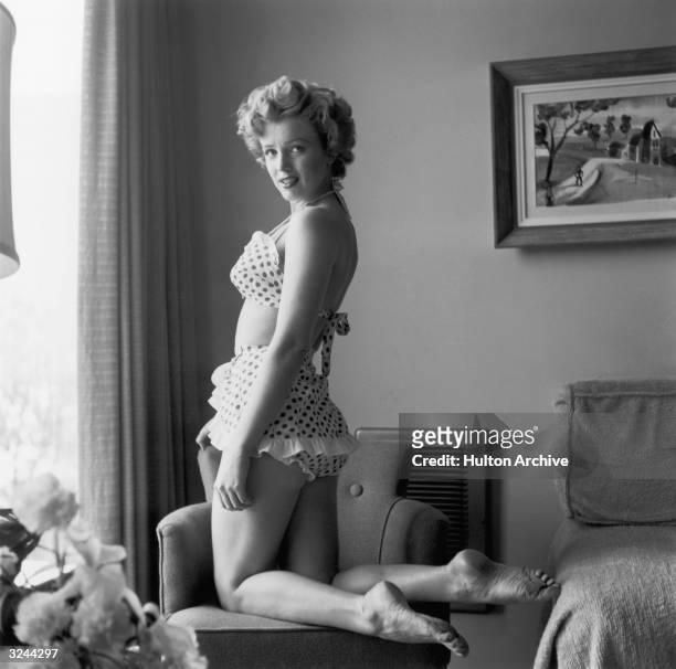 Portrait of American actor Marilyn Monroe kneeling in a chair in a two-piece polka dot bathing suit.