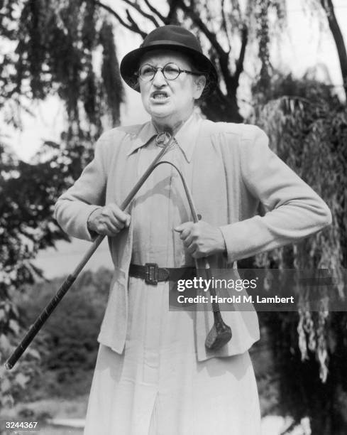 Middle-aged woman grimaces as she breaks a golf club with both hands while standing outdoors in a hat and glasses.