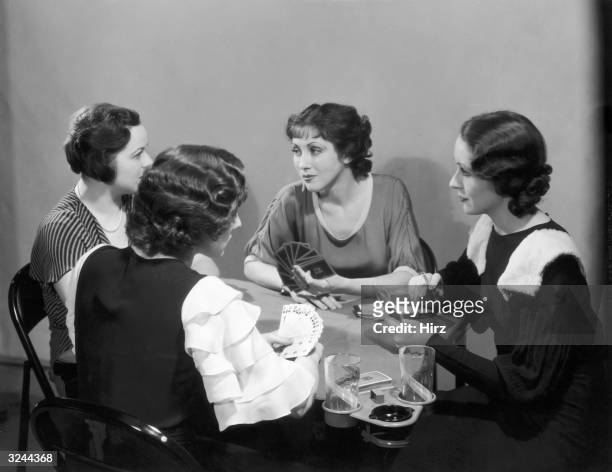 Four women in dresses play cards at a table with drink holders. One woman holds a cigarette and rests her hand on a scorepad.