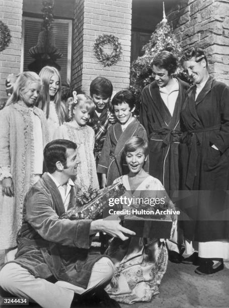 The cast of the television series 'The Brady Bunch' stands and watches American actors Robert Reed and Florence Henderson exchange wrapped Christmas...