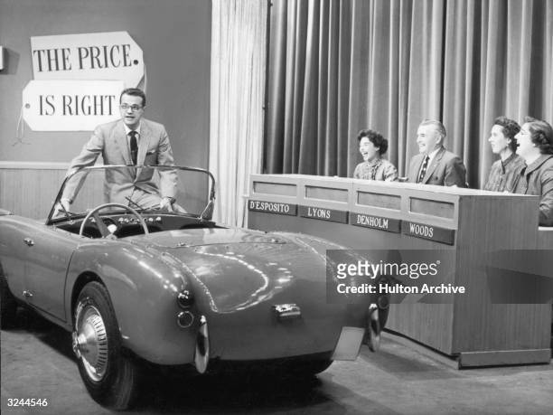 American TV host Bill Cullen sits on the edge of a convertible sports car as four contestants laugh in a still from the television game show 'The...