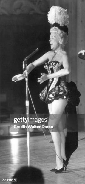 American actor Betty Grable sings on stage at a microphone, holding her hands outstretched and wearing a burlesque costume with a plumed cap, at the...