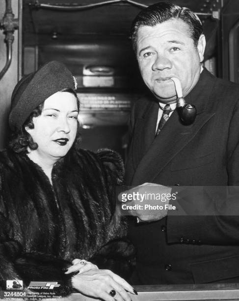 American baseball player George Herman 'Babe' Ruth smokes a pipe while wearing an overcoat and standing beside his wife, Claire Merritt Hodgson, who...