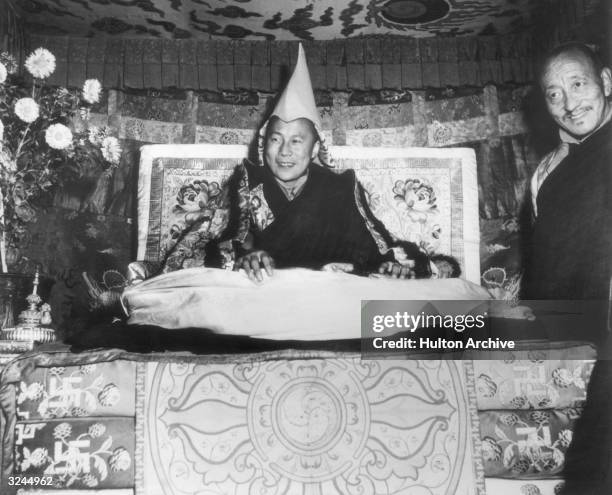 His Holiness the Dalai Lama, Tenzin Gyatso, seated on his throne and wearing the gold peaked cap which is his Crown, smiles while giving an audience...