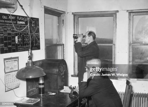 One elderly man sits with a telephone, as another elderly man stands and looks through binoculars, preparing for air raids at a civilian station,...