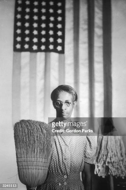 Ella Watson, a government charwoman, in Washington DC. She is wearing a polka dot smock, standing in front of an American flag, and holding a broom...