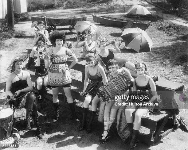 Group of young women wearing swimsuits relax on a beach, playing an accordion and horns in a promotional still for producer Mack Sennett's Bathing...