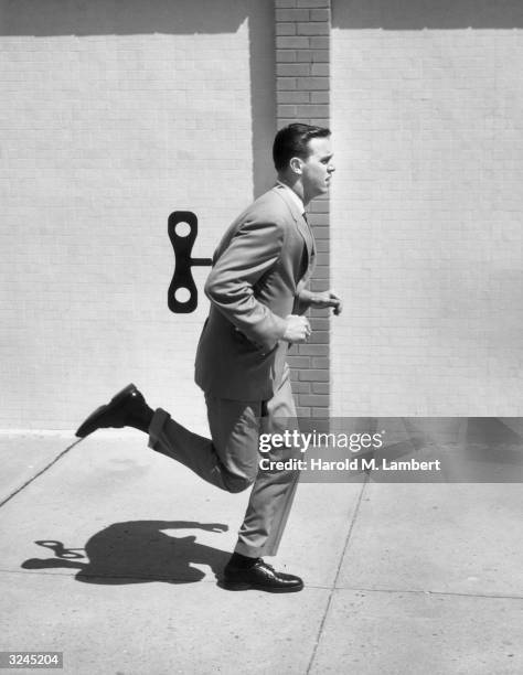 Businessman runs on the sidewalk with a wind-up toy key attached to his back, 1950s.
