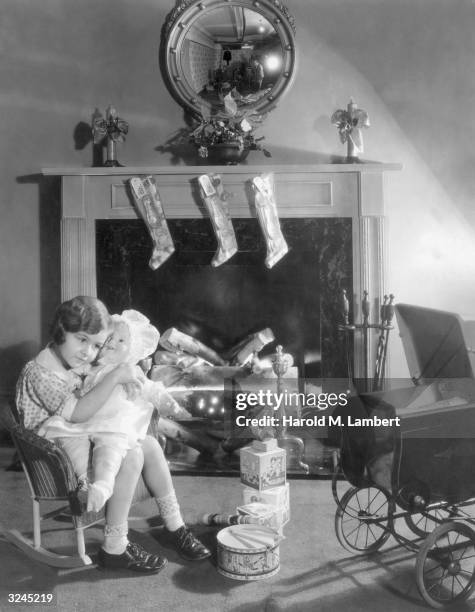 Young girl sits in a rocking chair near a fireplace hung with Christmas stockings and hugs a big doll. Other toys lie at her feet.