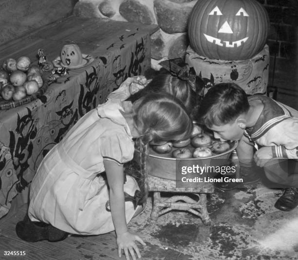 Young boy and girl crouch and bob for apples on Halloween, with an illuminated Jack-O-Lantern behind them, circa 1935.