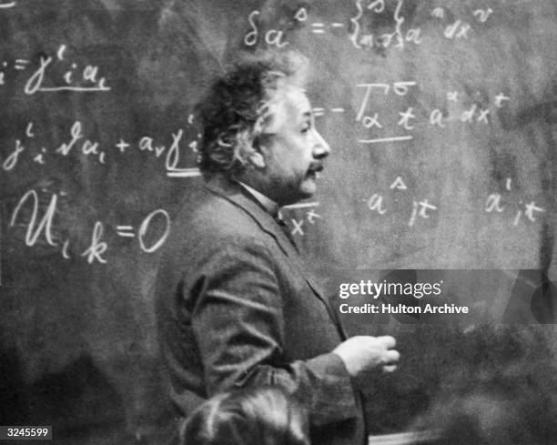 German-born physicist Albert Einstein standing beside a blackboard with chalk-marked mathematical calculations written across it.
