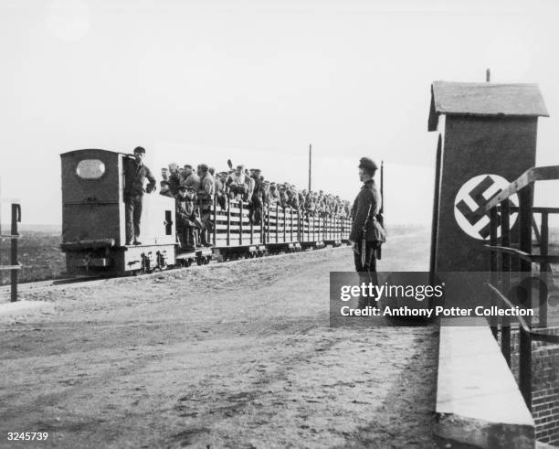 An armed Nazi sentry stands guard as a train full of people enters the gate of a concentration camp during World War II.