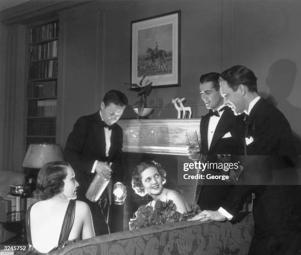 Two men and two women chat by the fireplace in a living room as a another man pours cocktails at a formal gathering.