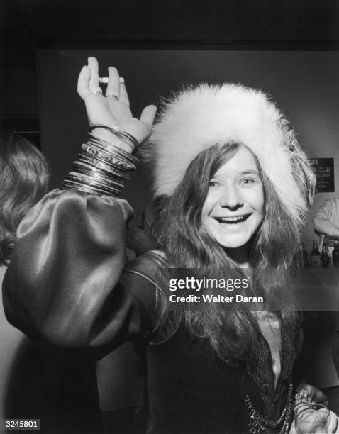 American rock singer Janis Joplin smiles and waves with a cigarette in one hand and a drink in the other while attending a party. She wears a fur hat.