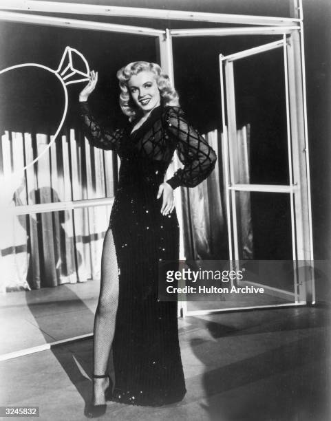 Full-length portrait of American actor Marilyn Monroe wearing an evening dress with a high slit revealing a fishnet stocking, standing in front of a...