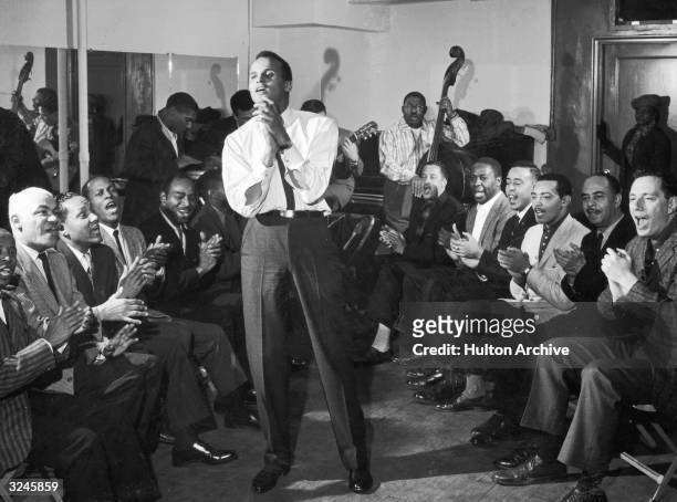 American singer and musician Harry Belafonte Jr. Sings and claps his hands along with a group of men sitting around him. A band plays in the back of...