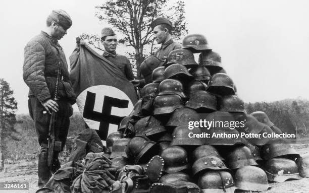Three Soviet soldiers display a Nazi flag and a pile of military helmets and boots, captured after a German Luftwaffe Field Regiment was destroyed...