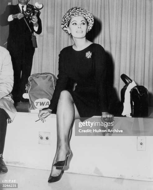 Italian actor Sophia Loren wears a knee-length black dress and leopard print hat while smoking a cigarette in an airport lounge. She sits next to a...