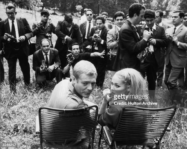 Italian actor Marcello Mastroianni and Swiss actor Ursula Andress sit outdoors while photographers take their picture on the set of director Elio...