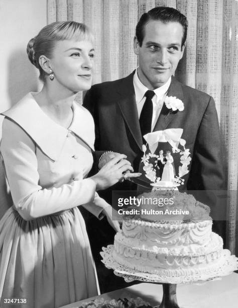 American actors Joanne Woodward, wearing a pale-colored dress with a pleated skirt, and Paul Newman, wearing a suit and tie, holding a knife together...