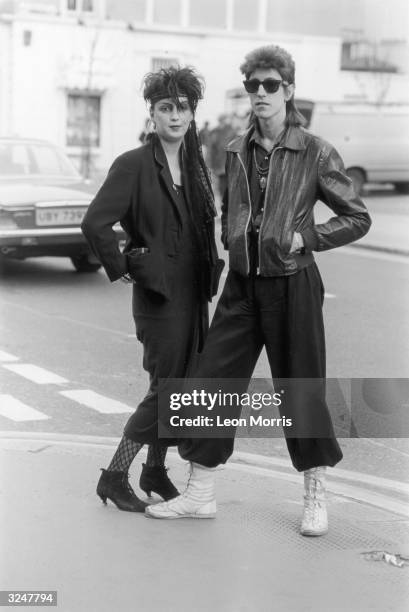 Couple with eighties hairstyles and outfits. The woman wears all black with heeled ankle boots and lace tights, he is wearing wide trousers and has a...