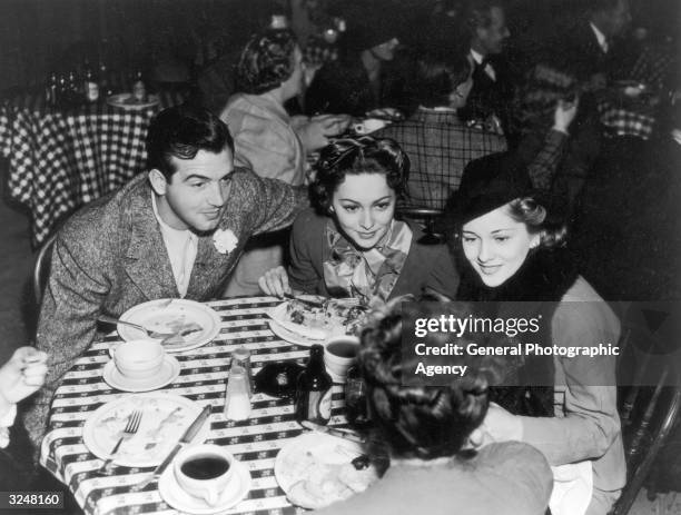 Olivia De Havilland leans in to hear a conversation while lunching with her sister Joan Fontaine and actor John Payne.