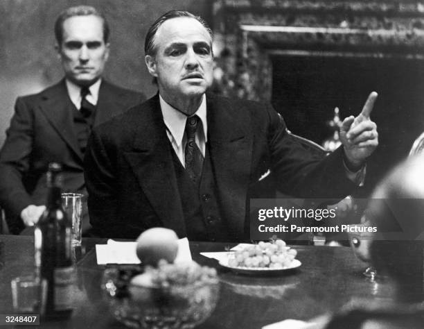 American actor Marlon Brando gestures at a table while American actor Robert Duvall sits behind him in a still from the film, 'The Godfather,'...