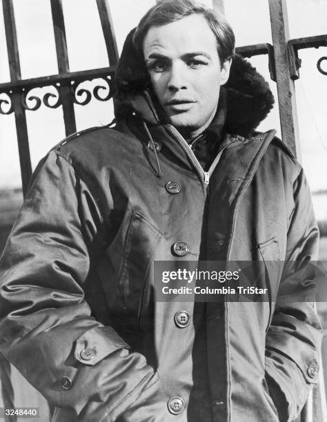 American actor Marlon Brando wears a parka and leans against an iron fence on the set of the film 'On the Waterfront,' directed by Elia Kazan.