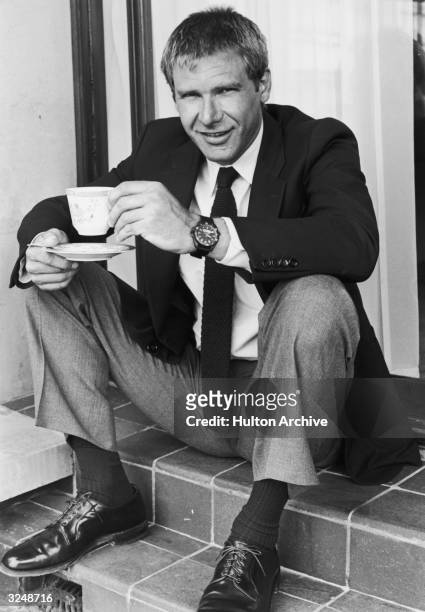 American actor Harrison Ford sits on steps, smiling while holding a teacup and saucer.