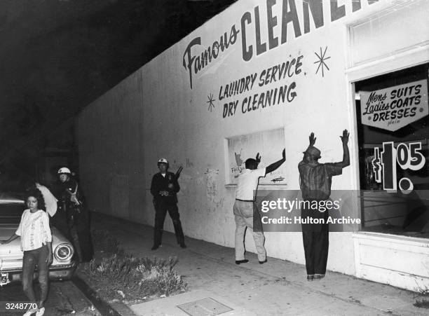 Two African-American men hold their hands up against the wall of a dry cleaners while being arrested by Caucasian state troopers during the Watts...