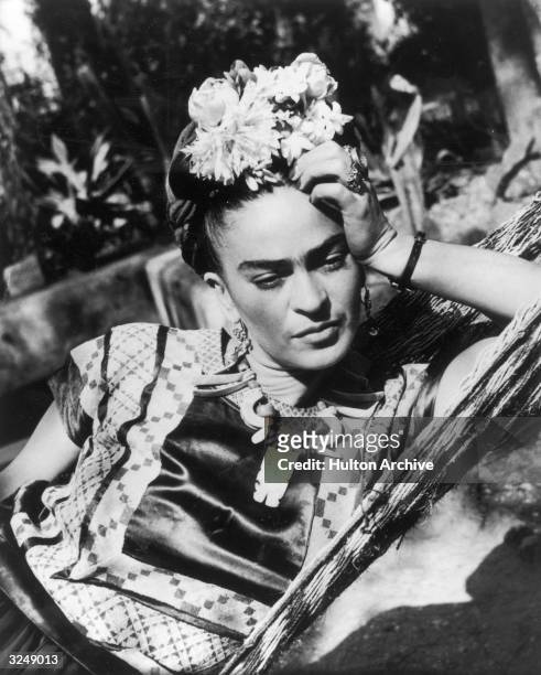 Mexican artist Frida Kahlo , wearing a folk costume and flowers in her hair, leans her head on her hand while lying in a hammock.