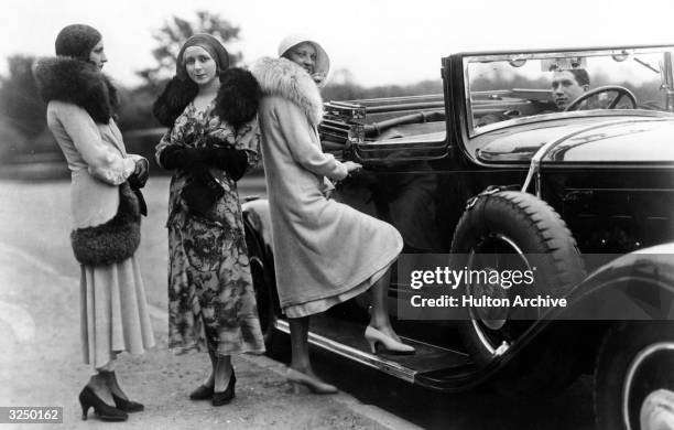 Women standing by a convertible car, wearing fur lined coats.