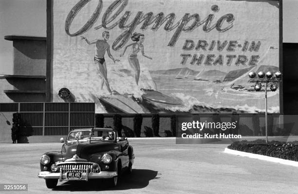 Couple in an open-top car driving past a sign advertising Hollywood's Olympic drive-in theatre in Los Angeles, California. Original Publication:...