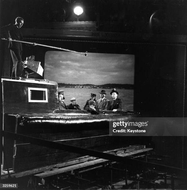 The background of an outdoor setting is projected onto a screen at Denham Studios as the actors perform. Cast members include Edward Lexy, Ralph...