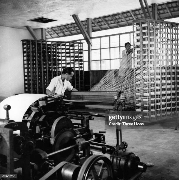 One worker at a textile factory in Bandung, Indonesia readies the warp threads on a weaving machine while the other steadies the bobbins of thread....