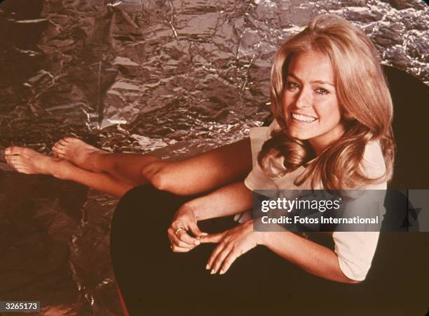 Portrait of American actress Farrah Fawcett dressed in a shirt as she sits and stretched her bare legs, early 1970s.