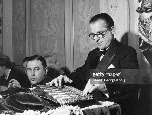 Guest temporarily suspends his drink to watch Anton Karas play the music he wrote for the film 'The Third Man' on his zither. Karas is sitting on his...