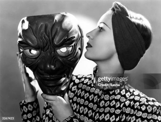 Valerie Hobson the British leading lady posing in profile and holding a large grotesque face mask in her hands.