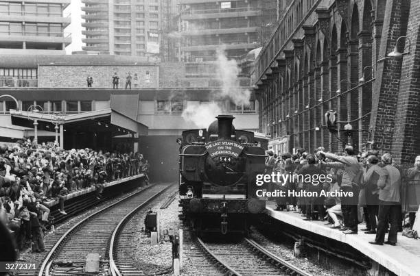 Steam train arrives at a station to a huge crowd. The modern towers of the Barbican Centre stand in the background.