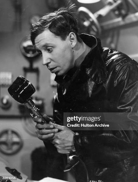 Waterproof clad man, probably George Formby, in an engine room seems very apprehensive about a what appears to be a microphone he is holding.