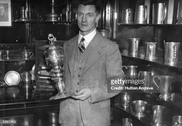 Man stands beside shelves of pewterware jugs and holds a pewter trophy.