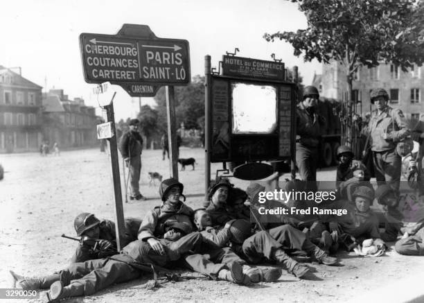 American paratroopers and glider troops resting in front of signs pointing to Cherbourg and Paris.