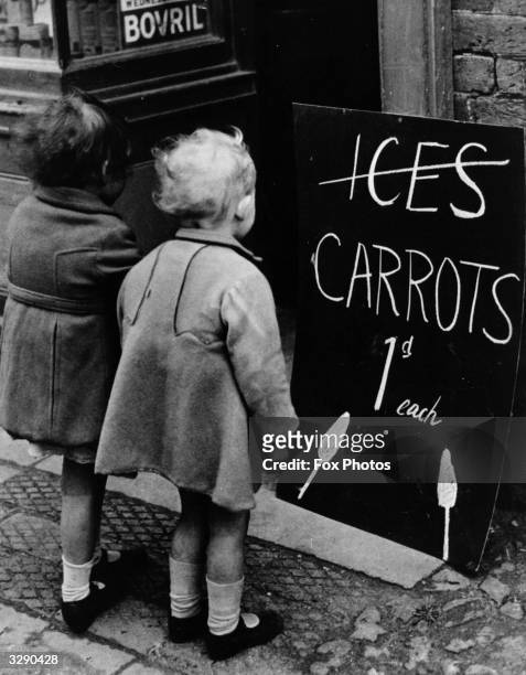 Two little girls reading a board advertising carrots instead of ice lollies. Wartime shortages of chocolate and ice cream made such substitutions a...
