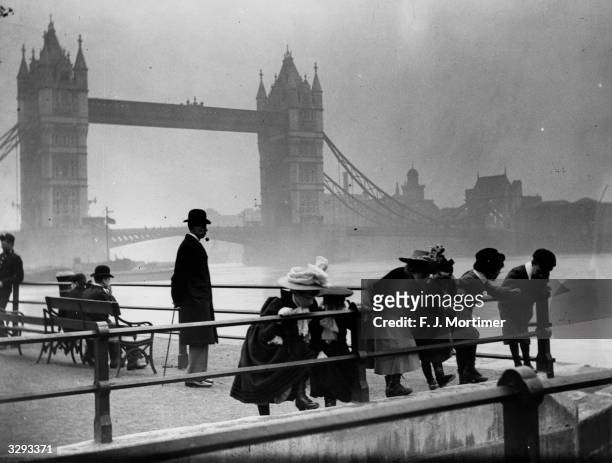 Children peer over the railings by the banks of the Thames with Tower Bridge in the background.